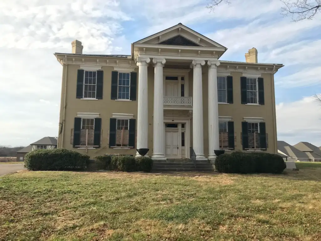 A large, two-story brick house with four tall white columns at the front entrance. The building has green shutters on the windows and is surrounded by a grassy lawn. The sky is clear with some clouds. Other houses are visible in the background.