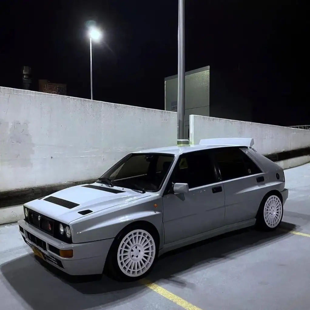 A gray, vintage hatchback car with white wheels is parked in a dimly lit outdoor parking lot at night. The car is positioned angled to the left, with a streetlight and concrete walls in the background.