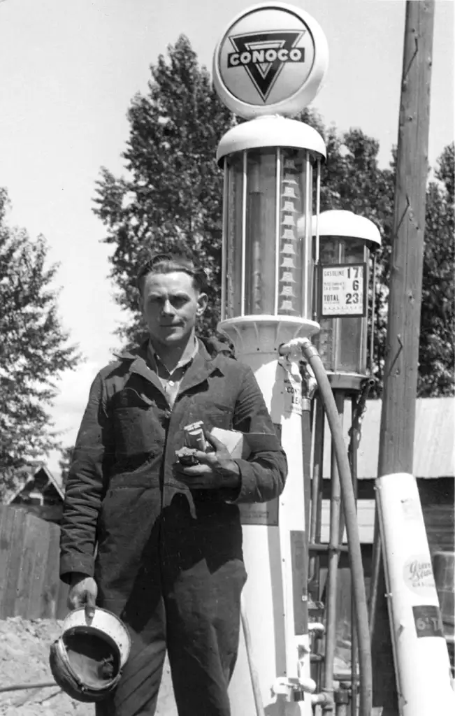 A man in a jumpsuit stands beside a vintage Conoco gas pump, holding a helmet and a rag. The pump displays old fuel prices, and the background has trees, a wooden fence, and a small building. The image is in black and white.