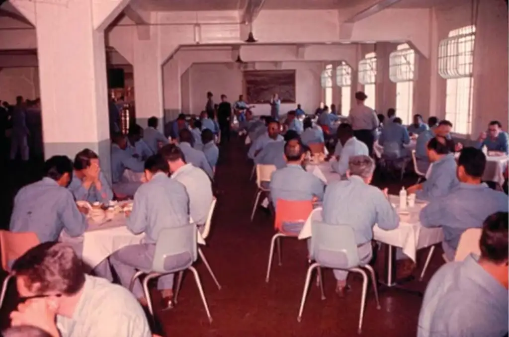 A large hall filled with men wearing identical blue uniforms, seated at rectangular tables. The room has large windows and fluorescent lighting. The setting appears to be institutional, with people eating and conversing.