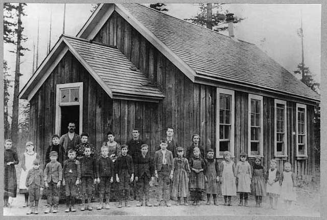 A group of children, along with a few adults, stands in front of a small wooden schoolhouse. The building features multiple windows and a steep roof. The background shows trees and an overcast sky, suggesting a rural setting.