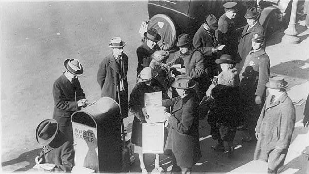 A black-and-white photo of people gathered around a mailbox on a sidewalk. They are wearing 1920s-style clothing, including coats and hats. A waste paper bin is visible, and a vehicle is parked nearby. Some individuals appear to be reading papers.