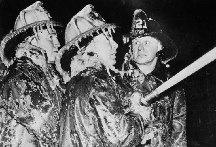 Three firefighters wearing ice-covered gear and helmets stand together in the cold. One holds a hose, focusing on their task. The icy conditions are evident on their uniforms, highlighting their challenging work environment.