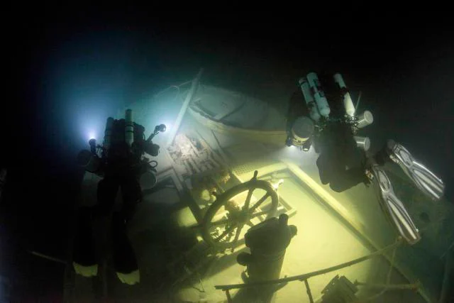 Two deep-sea divers examine a sunken shipwreck in dim light. They are equipped with scuba gear and torches, illuminating a ship's steering wheel and deck. The underwater scene is dark and mysterious, highlighting the remnants of the vessel.