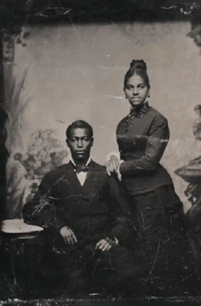 A vintage portrait of a Black couple from the 19th century. The man is seated, wearing a dark suit and bow tie, while the woman stands beside him, resting her hand on his shoulder, dressed in a dark, elegant gown. Both have serious expressions.