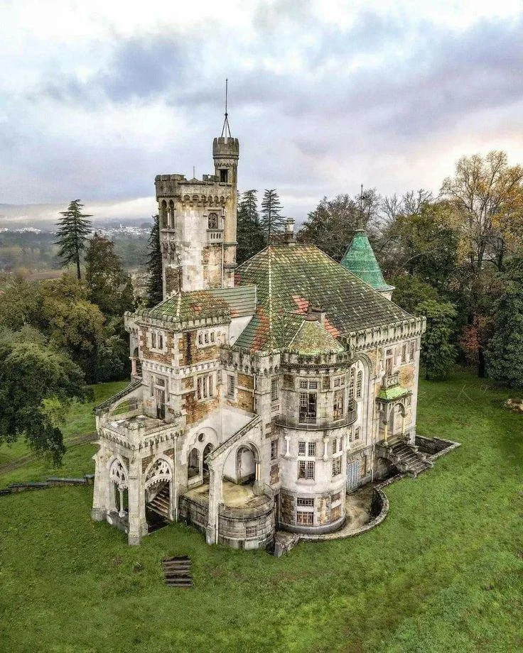 An ancient, weathered castle with a tall tower and arched entrances stands in a grassy landscape. Surrounded by trees, its green, mossy roof and intricate stonework reflect its age and history under a cloudy sky.