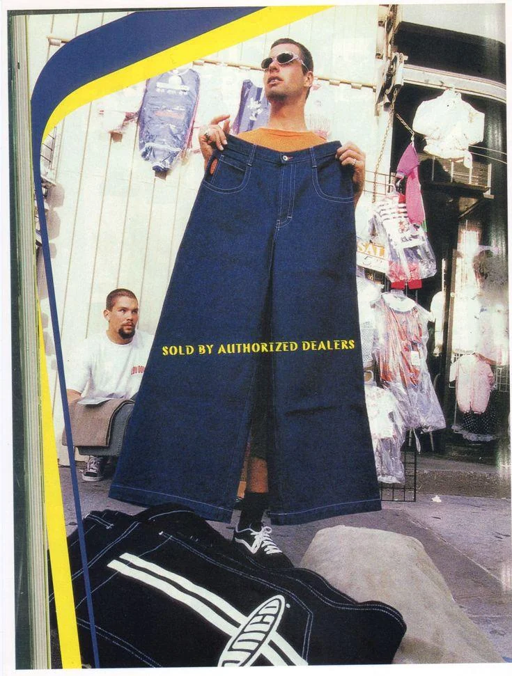 A man humorously stands holding an oversized pair of jeans in front of him on a busy street, with the text "SOLD BY AUTHORIZED DEALERS" displayed across the jeans. Another person sits in the background amidst hanging clothes.