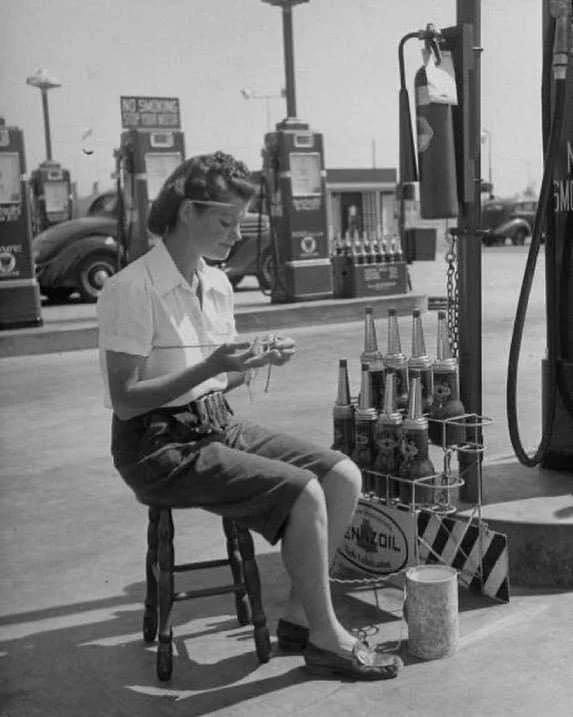 A woman in a white blouse and dark shorts sits on a stool at a vintage gas station, knitting. Around her are old gas pumps and oil cans. The setting has a mid-20th-century feel, with vintage cars in the background.