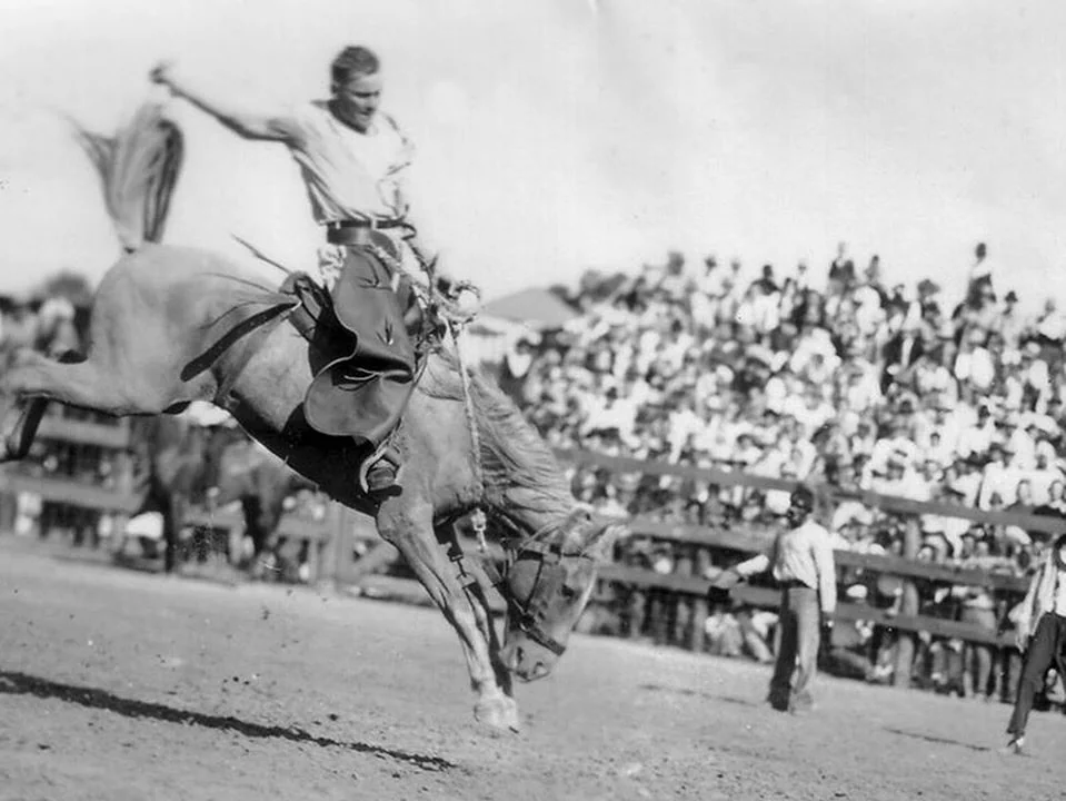 A cowboy rides a bucking horse at a rodeo event. He holds onto the reins with one hand, while waving his hat with the other. A crowd watches from the stands in the background. Dust is kicked up from the horse's hooves.