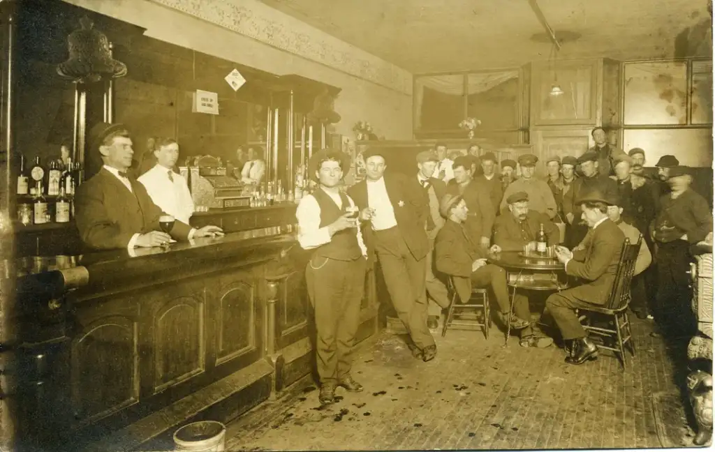 A sepia-toned image of a crowded bar scene from the early 1900s. Men in suits and hats stand at the bar and around a table, engaged in conversation. Bottles line the wooden bar counter, and a bartender stands behind it.