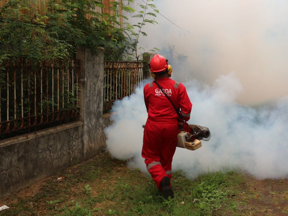 A person in red protective gear and a helmet is using a fogging machine to create smoke in an outdoor area next to a fence and greenery. The fog surrounds them as they work.