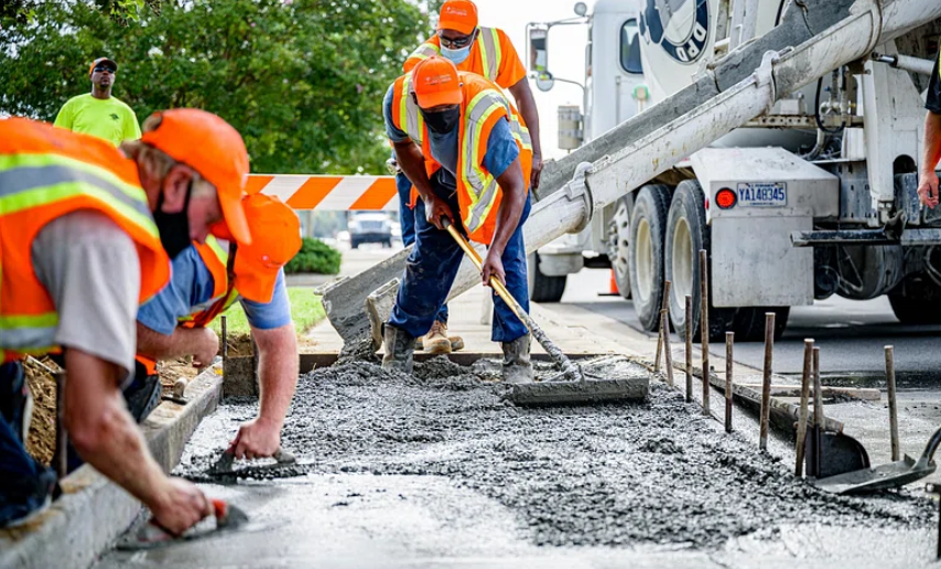 Workers in orange safety vests and helmets are smoothing wet concrete on a road. A cement truck is in the background, with a chute directing concrete onto the ground. Orange cones and a barricade are visible nearby.