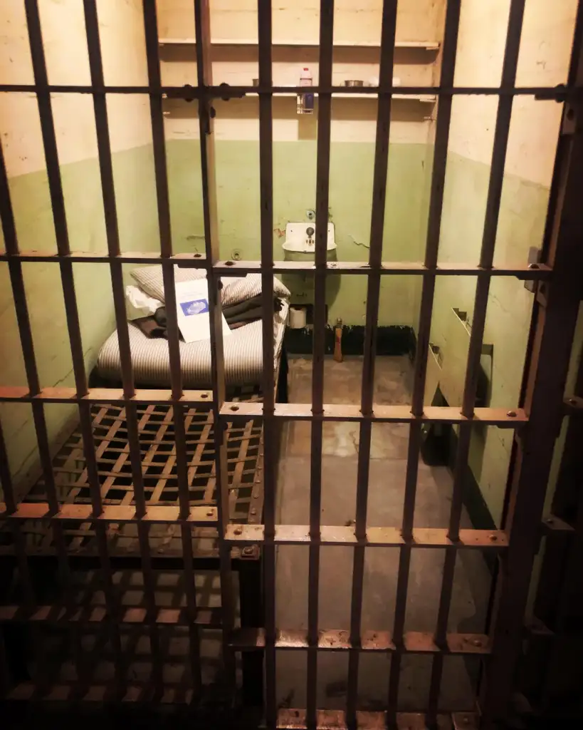 A dimly lit prison cell with metal bars in the foreground. Inside, there's a small bed with striped bedding, a sink, a toilet, and shelves on the wall. The walls are painted in a faded green and beige color.