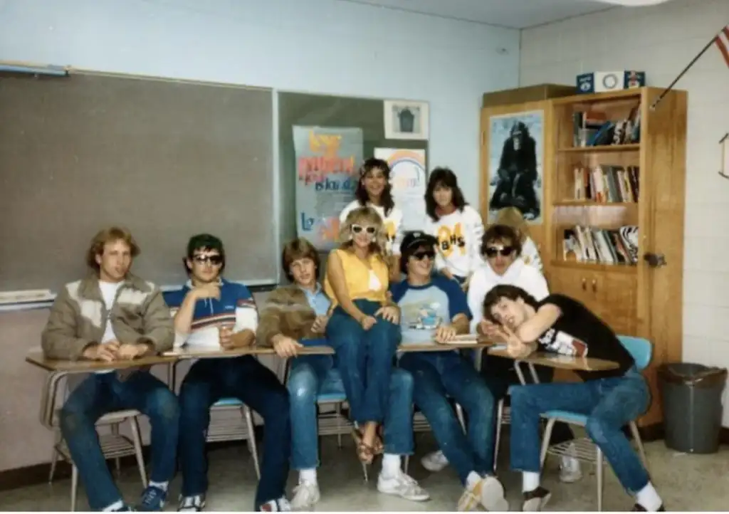 A group of nine people pose in a classroom, sitting and leaning on desks. They wear casual 1980s fashion, including sunglasses and jeans. Posters and a bookcase are visible in the background. The atmosphere is playful and relaxed.