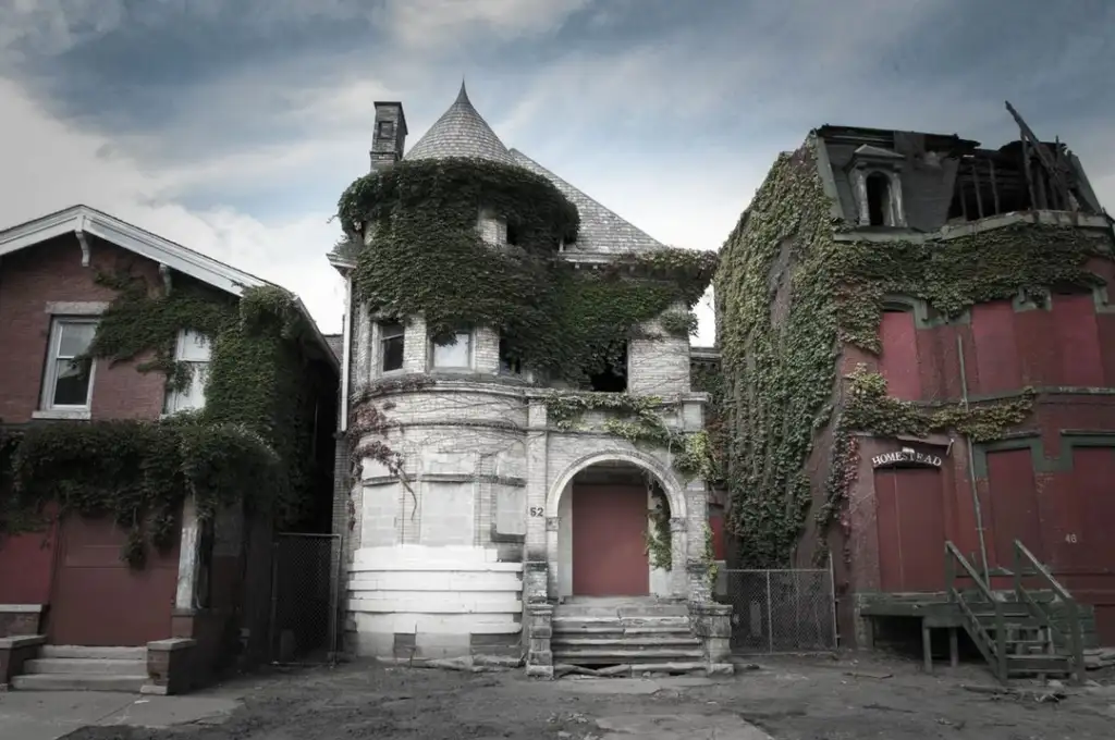 A group of abandoned brick buildings with boarded-up windows and doors, covered in thick ivy. The sky is overcast, giving a somber, eerie atmosphere.