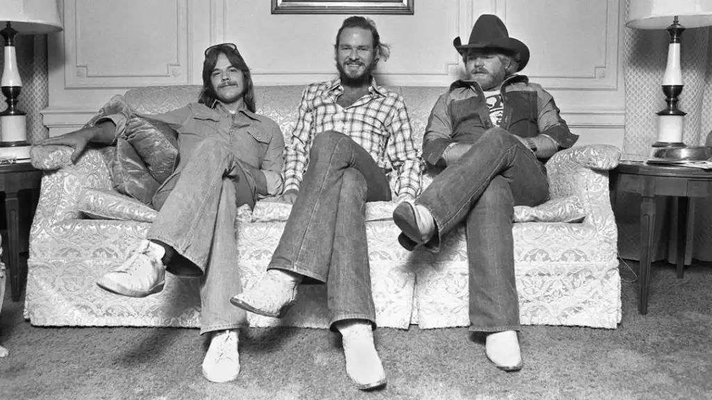 Three men sit on a patterned sofa in a living room. They are dressed in 1970s attire: bell-bottom pants and white shoes. The man on the right wears a cowboy hat. Two lamps and end tables flank the sofa against a paneled wall.