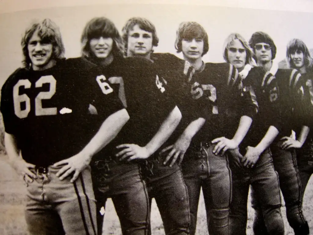 Seven young men stand in a row, wearing vintage football uniforms with numbers. They are posed with hands on hips, smiling. The image is in black and white, and the background is indistinct.