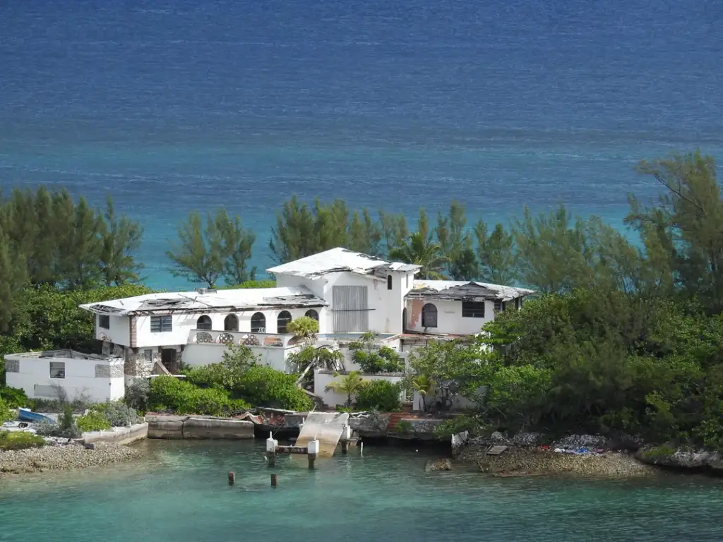 A white, abandoned beachfront house with a damaged roof surrounded by lush greenery. The property is next to a small dock, with clear turquoise waters and a backdrop of dark blue ocean in the distance.