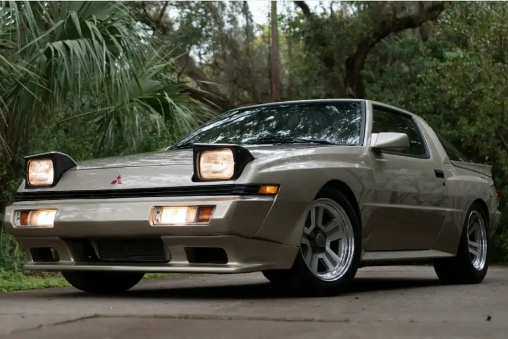 A beige classic Mitsubishi Starion car with pop-up headlights is parked outdoors on a concrete surface. It's surrounded by lush green foliage and trees, creating a vintage and natural setting.