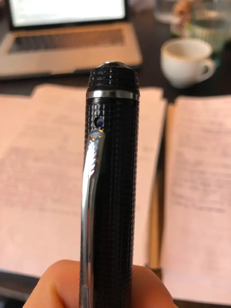 Close-up of a person holding a textured black pen with a silver clip. Blurred background shows handwritten notes on papers, an open laptop, and a white coffee cup on a desk.