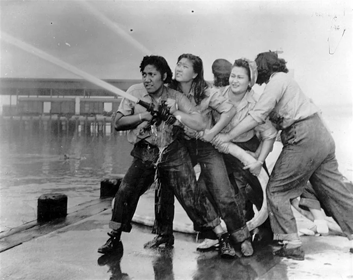 A group of five women, smiling and laughing, work together to direct a powerful stream of water from a hose on a wet dock. They are dressed in casual, practical clothing, suggesting they are involved in a firefighting or training activity.