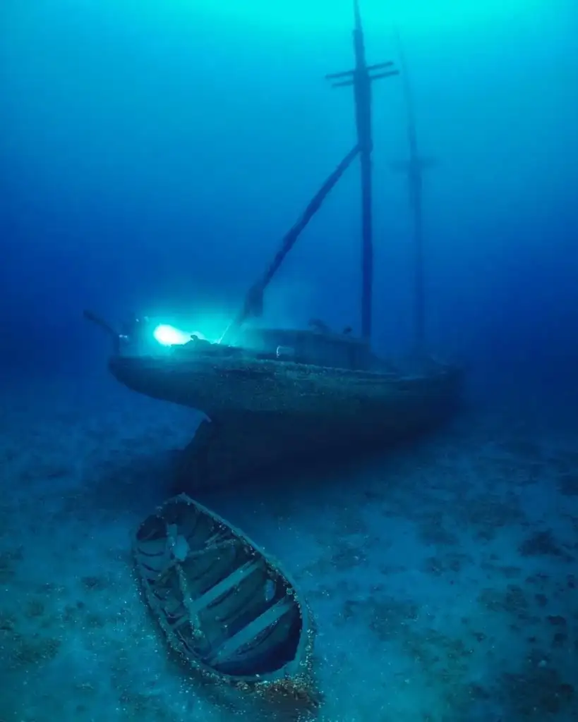 A sunken ship rests on a sandy ocean floor, illuminated by a blue light. Its masts reach upwards, surrounded by a hazy blue ambiance. A small wooden boat lies in front, partially buried in the sand.