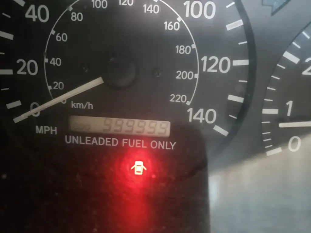 A car dashboard showing a speedometer in kilometers per hour and miles per hour. The odometer reads 999,999. Below, a red warning light shaped like a car engine is illuminated.