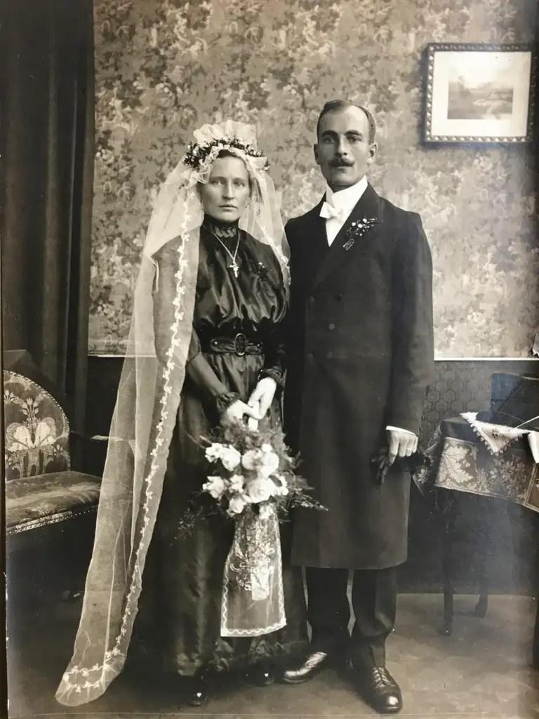 A black and white photo of a bride and groom. The bride wears a long veil and holds a bouquet, while the groom is in a formal suit with a bow tie. They stand in an ornate room with patterned wallpaper and a framed picture on the wall.