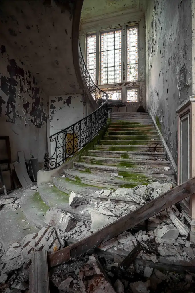 A dilapidated staircase in an abandoned building, with peeling paint and debris scattered on the steps. Moss covers parts of the steps. A large window with a grid pattern casts light onto the staircase, highlighting the decay.