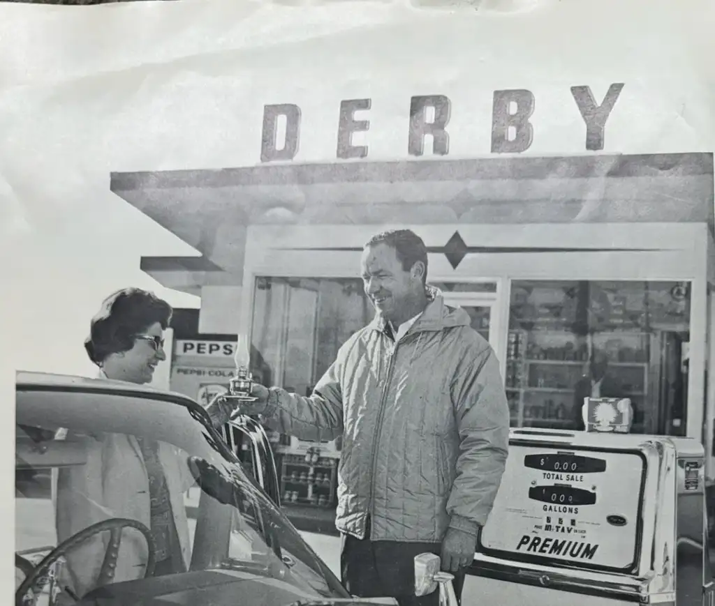 A vintage black-and-white photo shows a man in a jacket smiling as he hands a set of keys to a woman seated in a car. They are at a Derby gas station, with a retro pump displaying "PREMIUM" in the foreground.