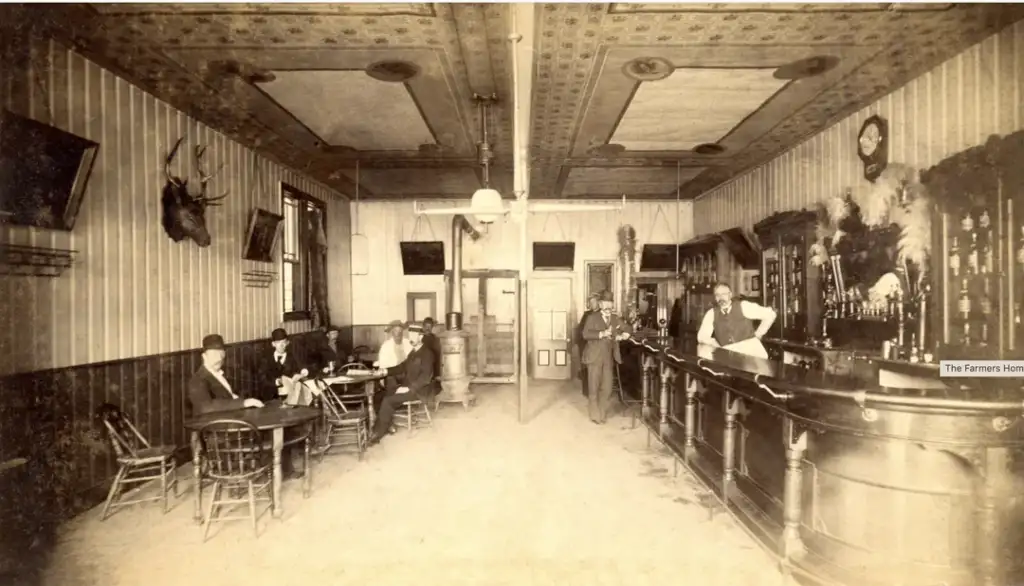 Sepia-toned photo of an old-fashioned saloon interior. Men in suits and hats sit at tables on the left, while others stand at a long bar on the right. Deer heads are mounted on the walls, and ornate ceilings are visible. Vintage decor throughout.