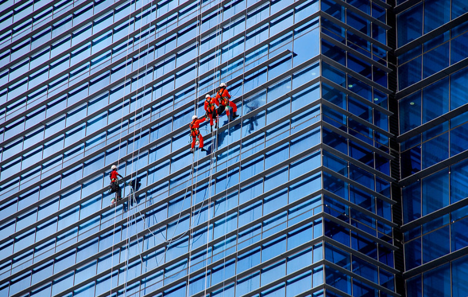 Workers in orange uniforms are suspended on ropes, cleaning the glass windows of a tall building with a grid-like pattern. One person is cleaning on the left, while a group works together in the center.