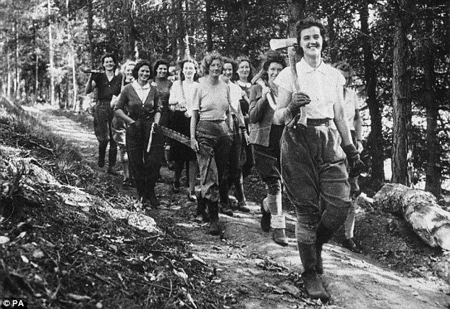 A group of women, dressed in work attire including trousers and boots, walk through a forested path. The leading woman carries an axe over her shoulder. Trees line the path, and they appear cheerful and determined.