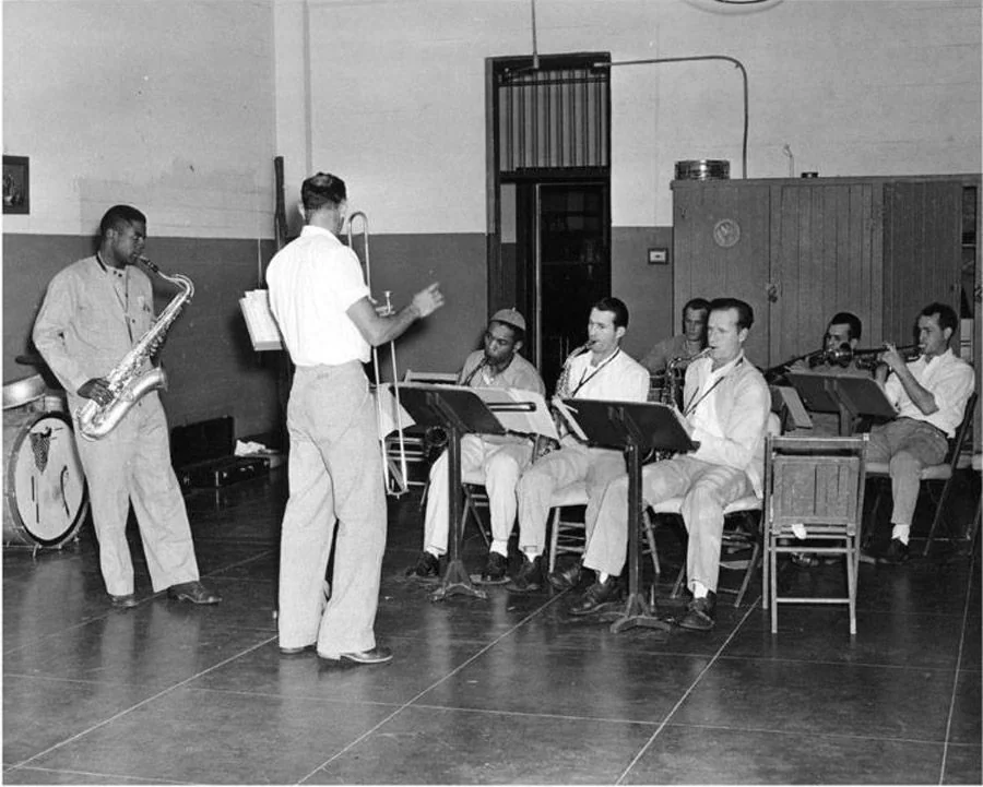 A group of musicians in a room, with a man conducting them. One musician plays a saxophone while others are seated playing various instruments. A drum set is visible in the background. All are dressed in light-colored clothing.