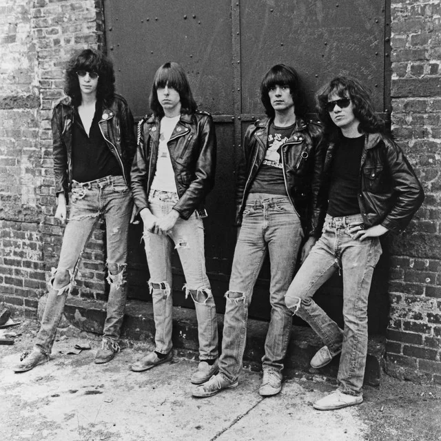 Four people stand against a brick wall, wearing leather jackets and ripped jeans. The setting has an urban, gritty feel, with a closed metal door behind them. They exude a punk rock style.