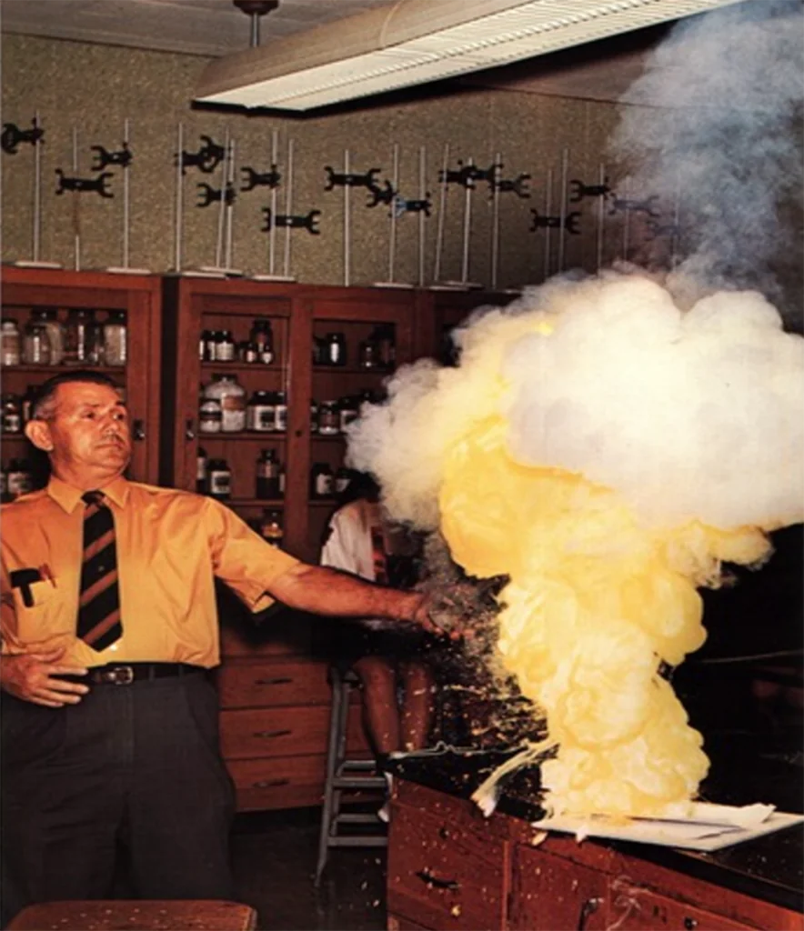 A man in an orange shirt and striped tie conducts a science experiment in a classroom. A chemical reaction produces a large cloud of yellow smoke or foam on a lab table. Shelves with bottles are visible in the background.