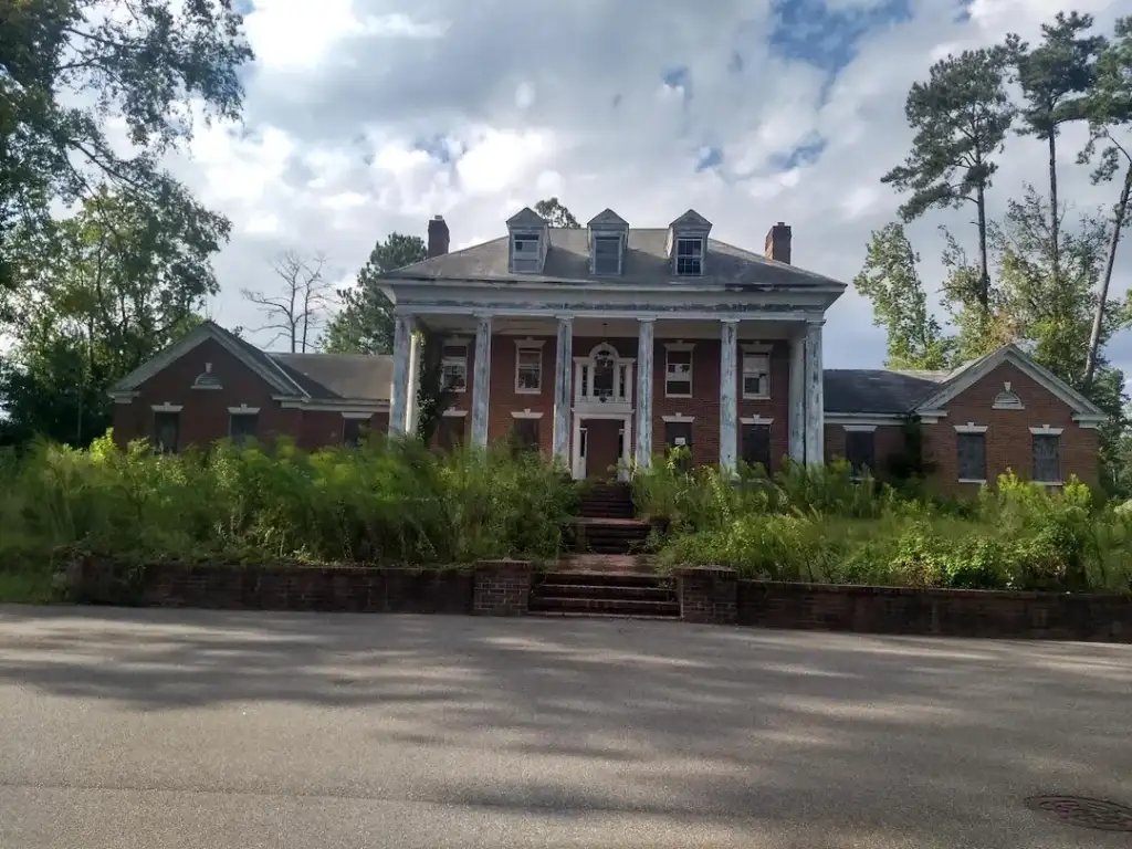 An old, weathered mansion with a brick facade and white columns stands amidst overgrown vegetation. The two-story building features a central staircase leading to the entrance. The sky is partly cloudy, and tall trees surround the property.