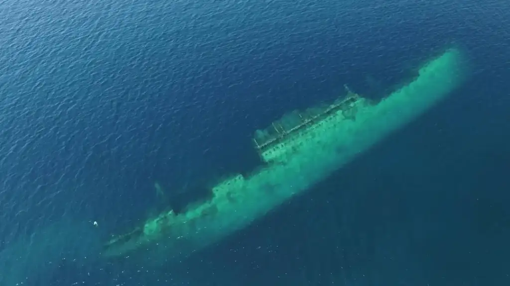 Aerial view of a large shipwreck submerged in clear, deep blue water. The outline of the ship is fully submerged but visible, with parts of the structure discernible beneath the surface, contrasting with the surrounding sea.