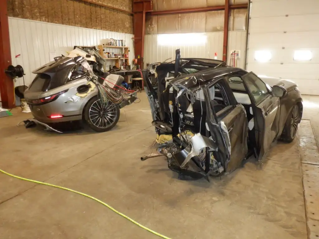 Two severely damaged cars are inside a workshop. The car on the right has significant rear damage, while the car on the left has a clutter of wires and equipment attached to its rear. The setting is industrial, with concrete floors and metal walls.