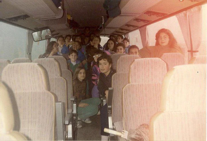 A group of people, mostly children, are seated inside a bus. The bus is half full, and the passengers are looking at the camera. The image has a vintage feel with dim lighting and a slight red tint.