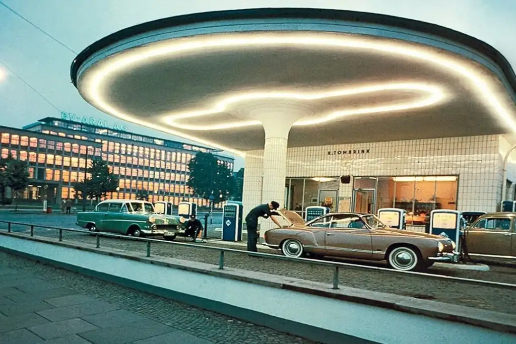 A retro gas station with a futuristic, circular canopy lit by neon lights. Two vintage cars, one green and one brown, are parked near the pumps. A person is checking under the hood of the brown car. Buildings are visible in the background.