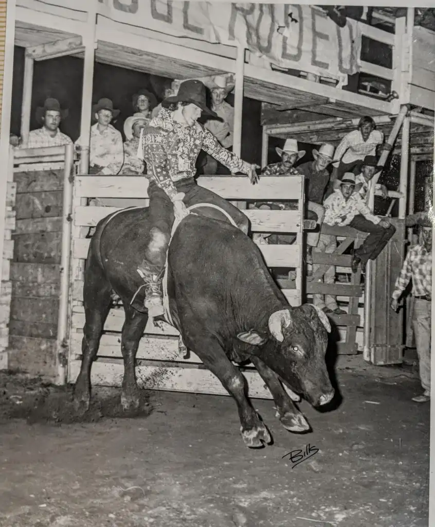 A black-and-white image of a rodeo scene. A rider wearing a hat and patterned shirt holds on tightly while riding a bucking bull in an arena. Spectators watch in the background, some sitting on fences. A "Rodeo" sign hangs overhead.