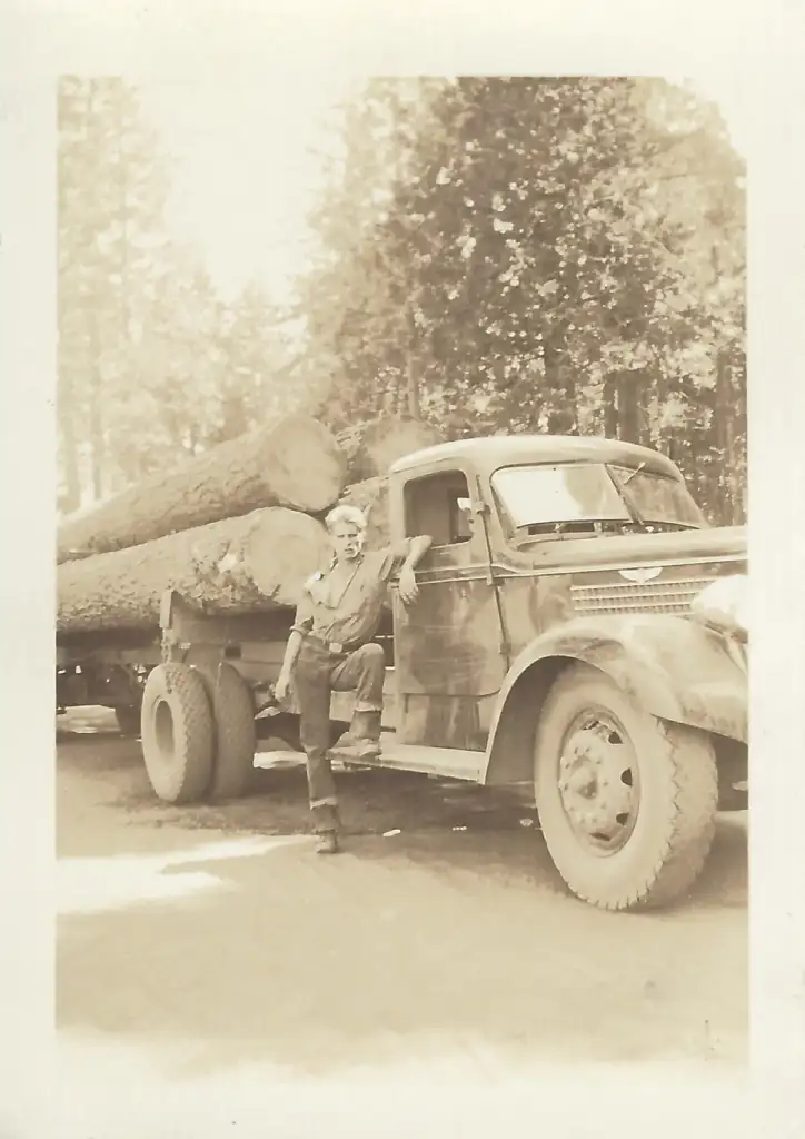 Sepia-toned image of a person leaning on a vintage logging truck loaded with large logs. The truck is parked on a forest road, with tall trees in the background, evoking a historical or rustic setting.