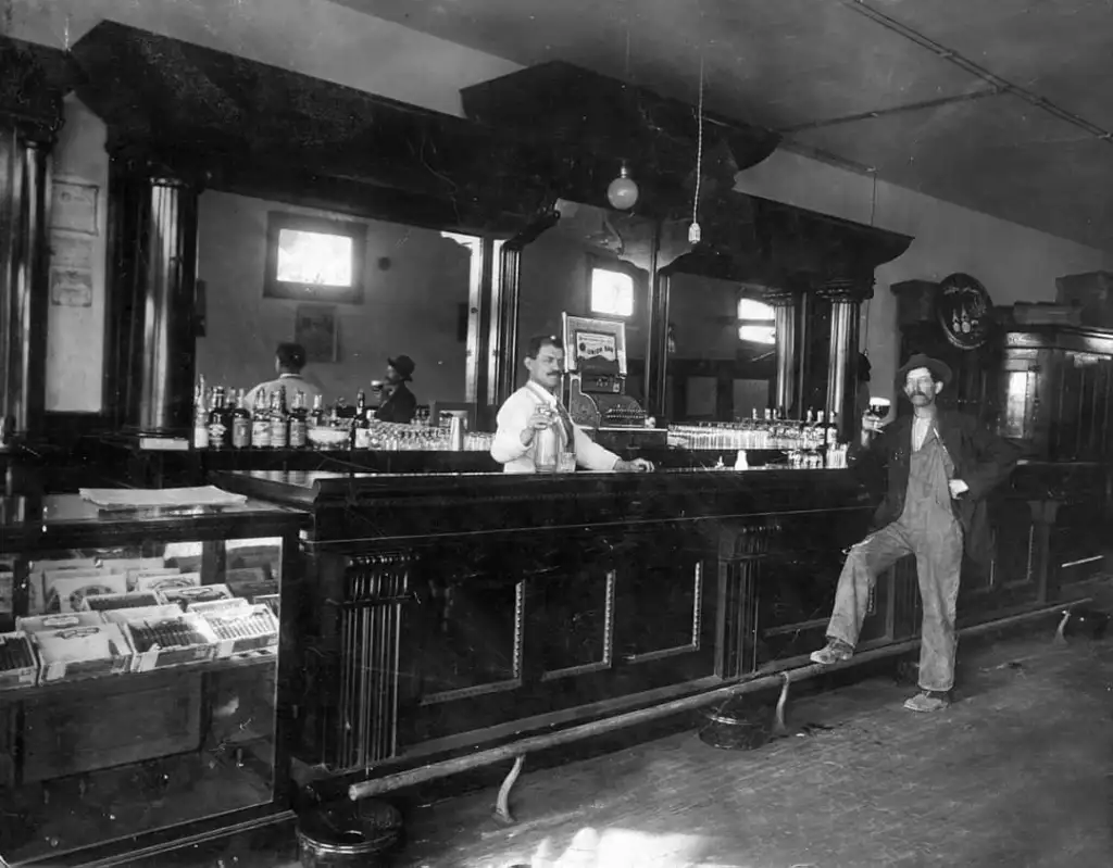 A vintage black-and-white photo of a bar scene. A bartender stands behind a long wooden counter lined with bottles and a cash register, while a man leans against it. The room features ornate woodwork and a high ceiling.