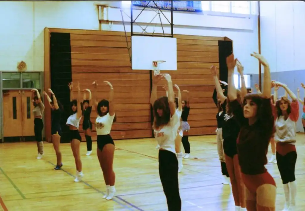 A group of women in workout attire performs a synchronized dance or exercise routine in a gymnasium. They have their arms raised and are standing in rows on the wooden floor, with a basketball hoop and a large padded wall visible in the background.