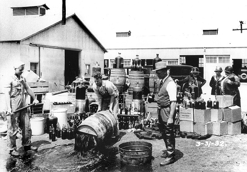 A group of men, including police officers, are gathered around a barrel outdoors. One man is pouring liquid from the barrel onto the ground, with numerous bottles and boxes in the background. The scene appears to depict a prohibition-era alcohol disposal.