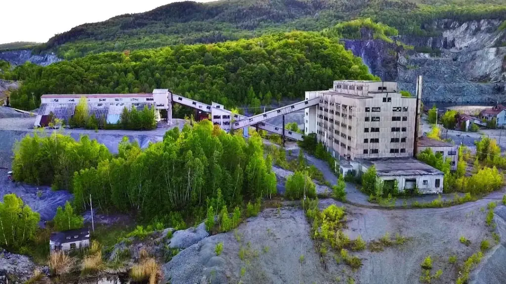 Aerial view of an abandoned industrial complex with large, aged buildings surrounded by lush green trees and hills. The structures appear weathered, with connecting walkways and overgrown vegetation around the site.