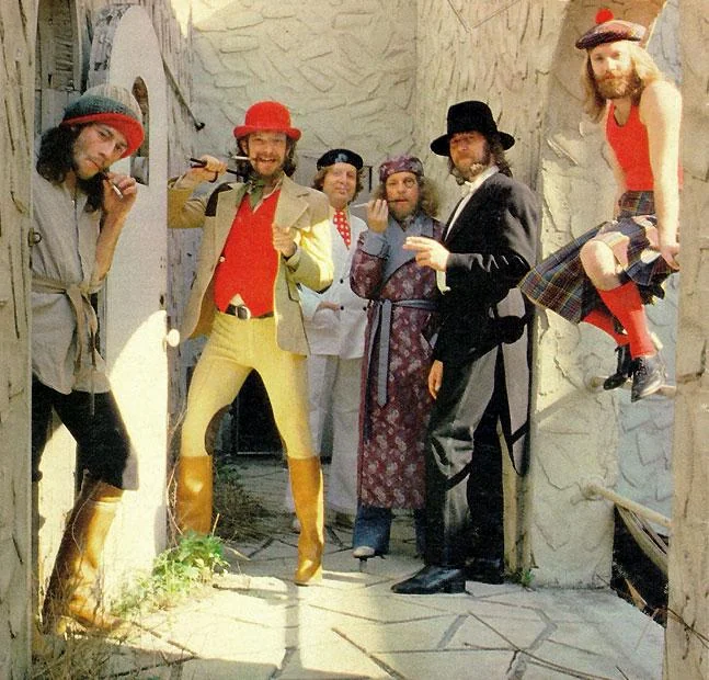 A group of six people in colorful, eclectic costumes poses in a stylized, textured outdoor setting. One person wears a red outfit with a hat, and another dons a kilt. The mood is playful and creative.