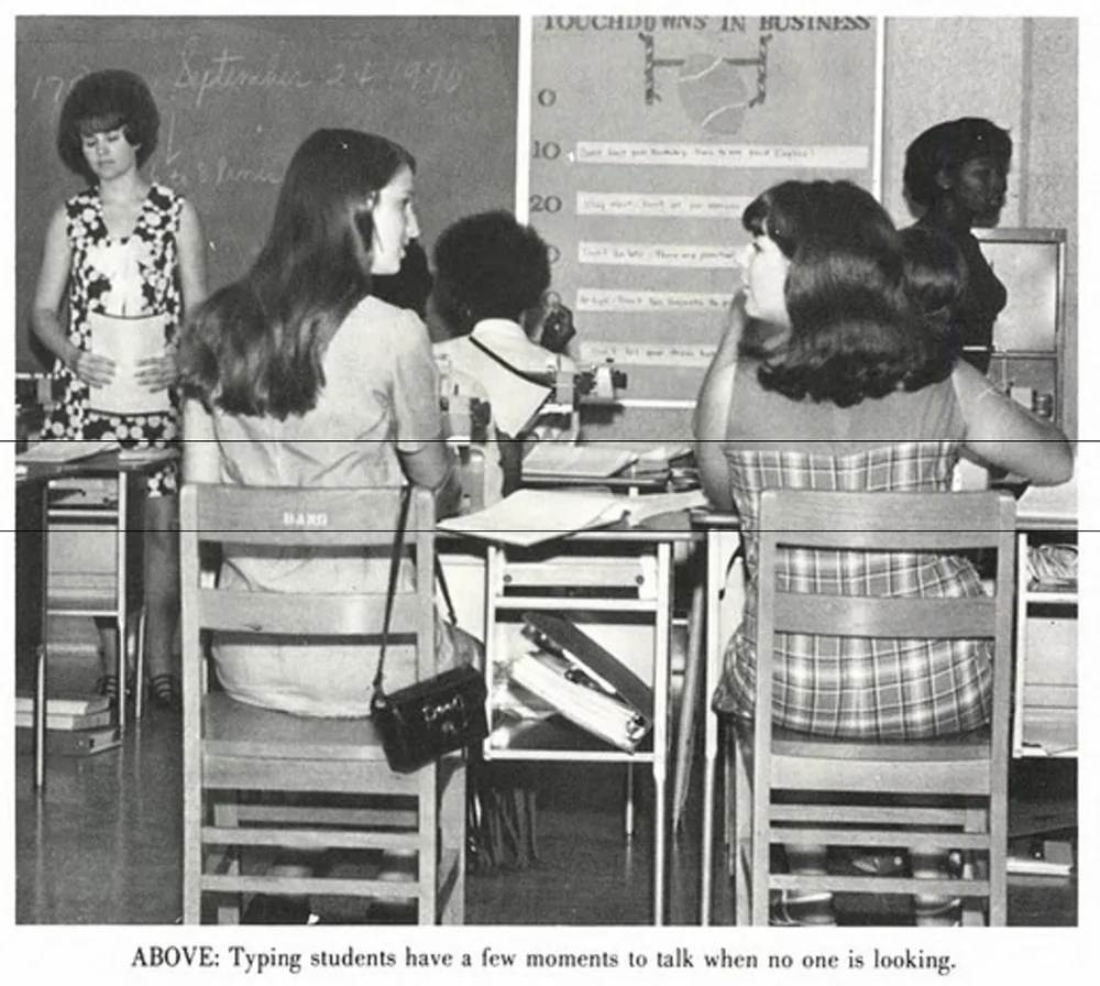 A classroom scene with several students sitting at desks. They are engaged in conversation with one another. The chalkboard in the background has writing on it, and there's a poster titled "Touchdowns in Business." A teacher stands nearby.