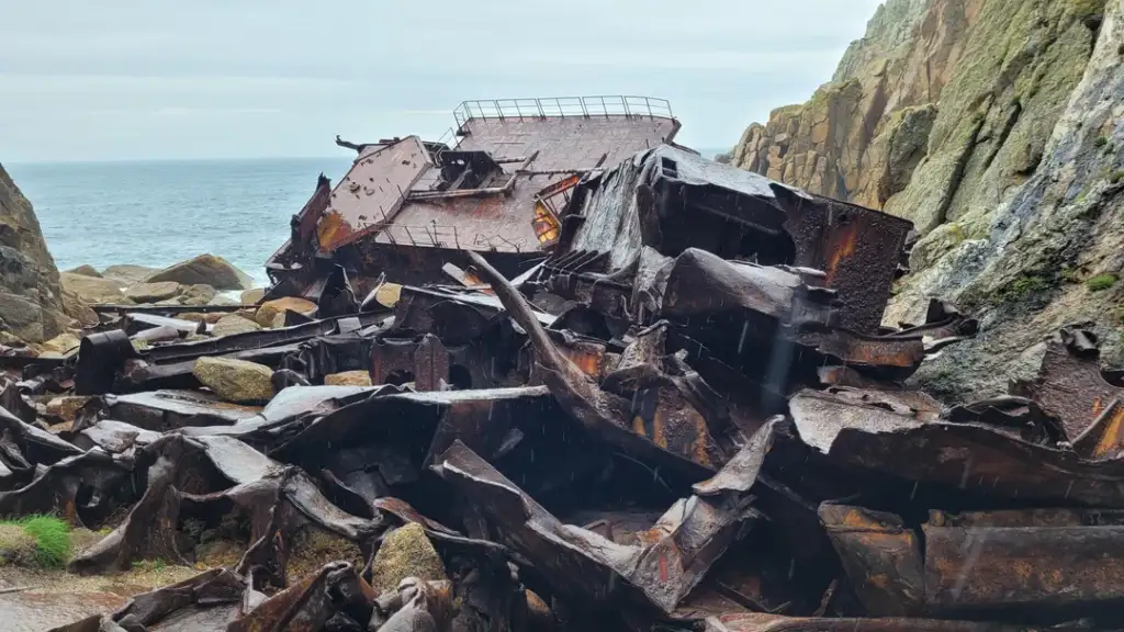 Rusty remains of a large, wrecked ship lie scattered on a rocky shore. The metal is twisted and corroded, surrounded by jagged rocks. The ocean is visible in the background beneath a cloudy sky.